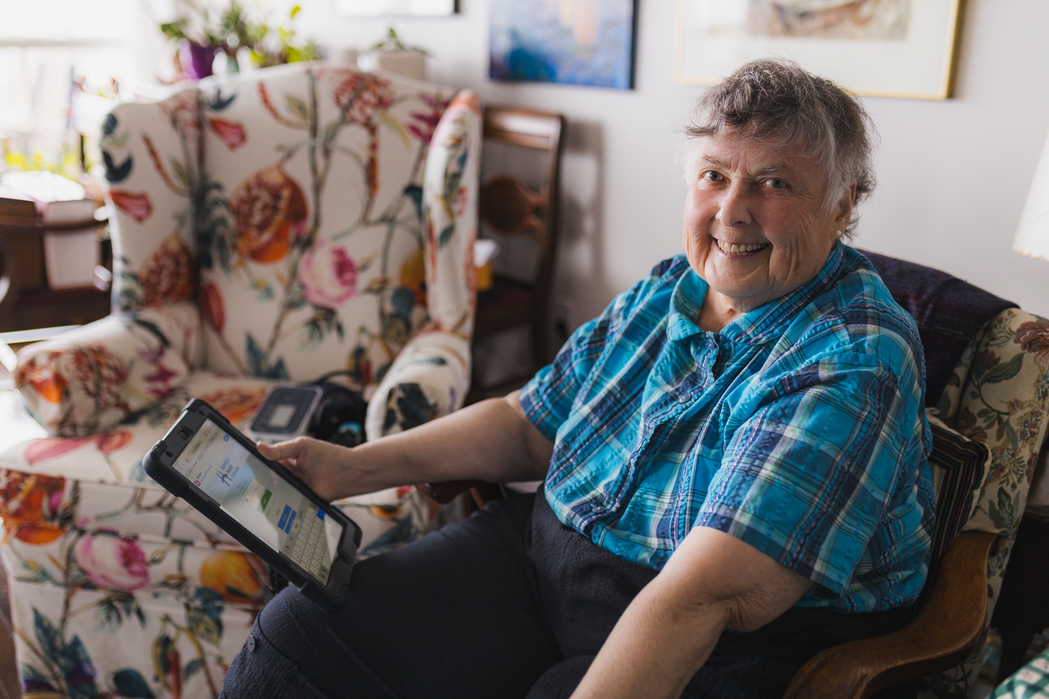 Peggy Kelly sits in a chair in her living room, holding an ipad. A blood pressure cuff is on another chair in the background.